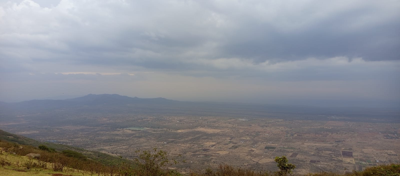A vast, panoramic view from a high vantage point overlooking a dry, flat valley. The landscape is dotted with small settlements and farms, all under a heavy, overcast sky.